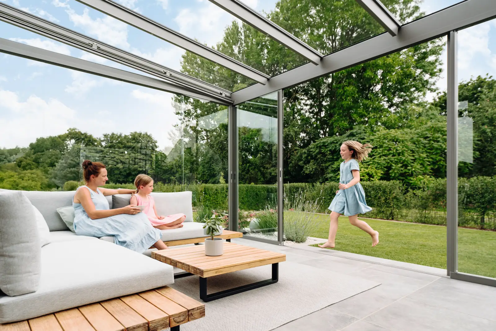 Mutter und Tochter sitzen auf dem Sofa im Glashaus, ein Mädchen läuft durch den Garten heran