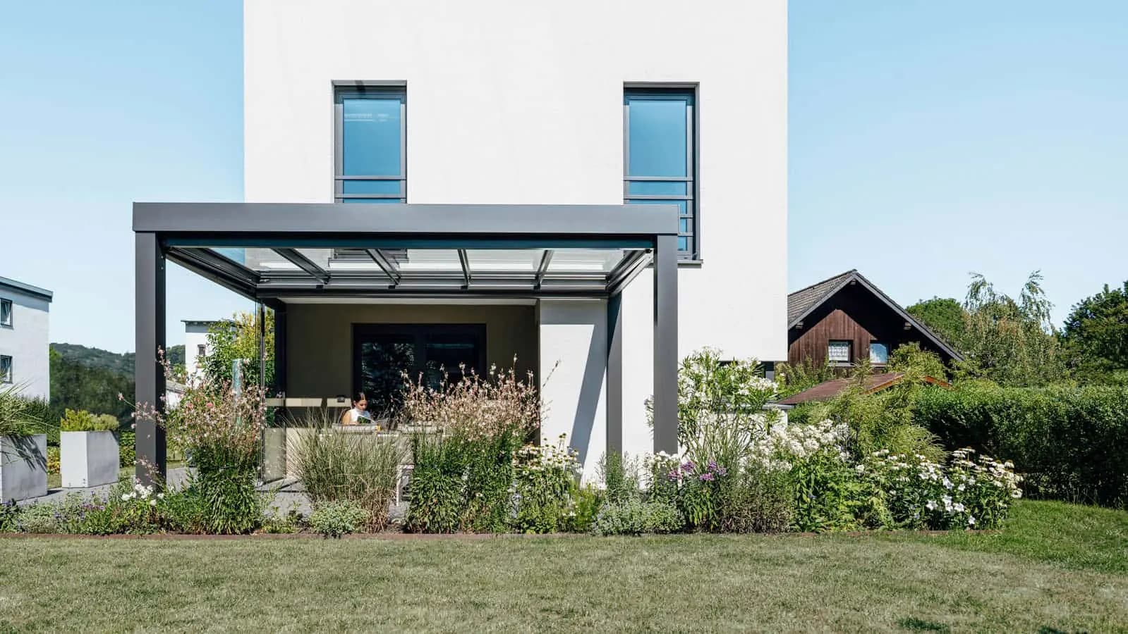 Frau liest unter einer anthrazitfarbenen Terrassenüberdachung am modernen Einfamilienhaus
