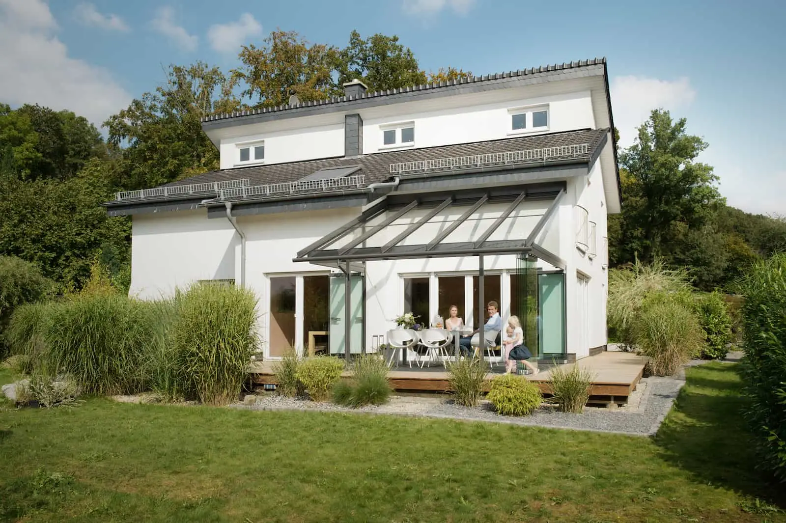 Familie sitzt am Esstisch unter einer Glas-Terrassenüberdachung am Einfamilienhaus