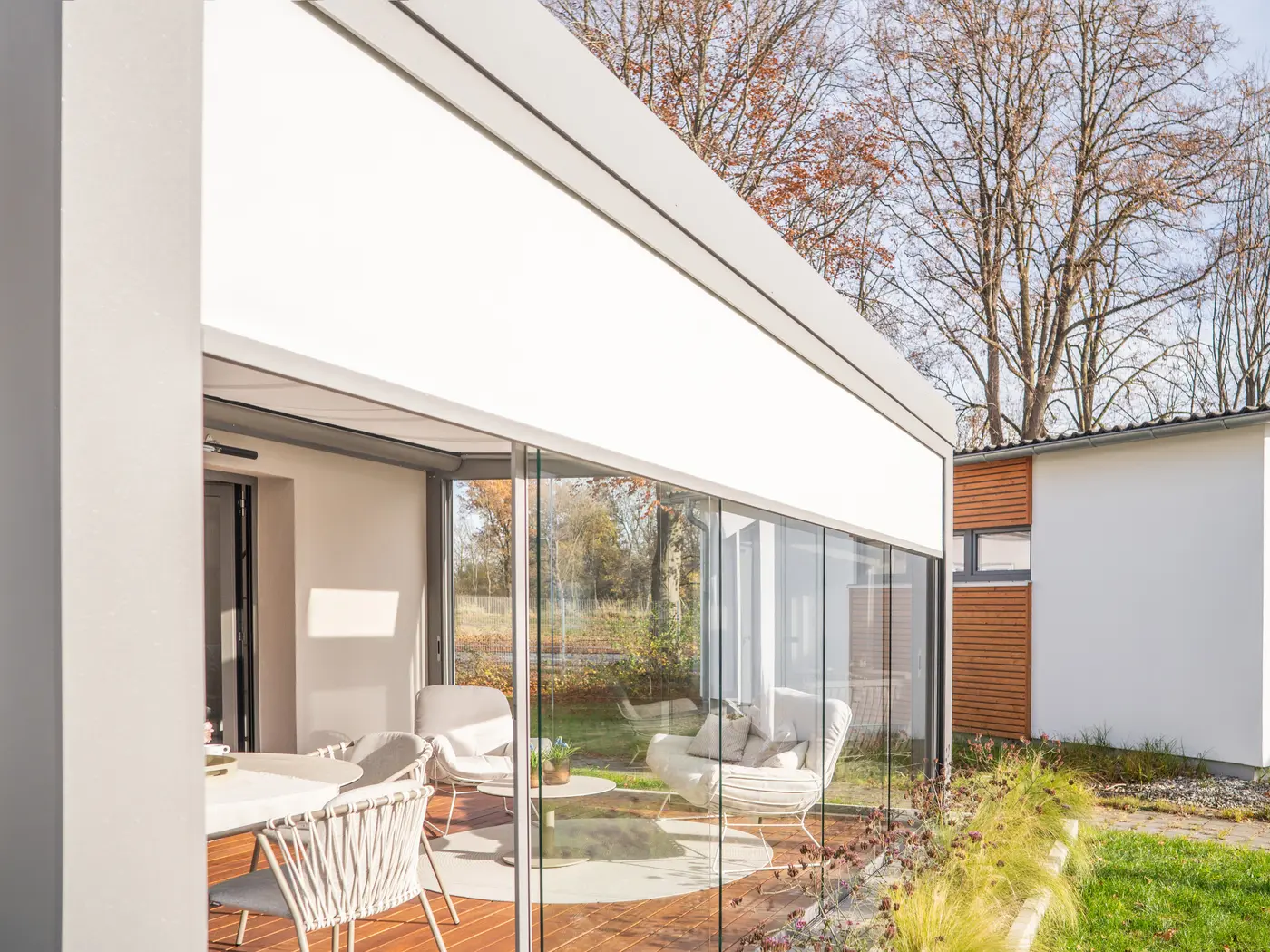 Seitlich heruntergelassene Senkrechtmarkise an der Glasfront, davor der Garten im Herbst