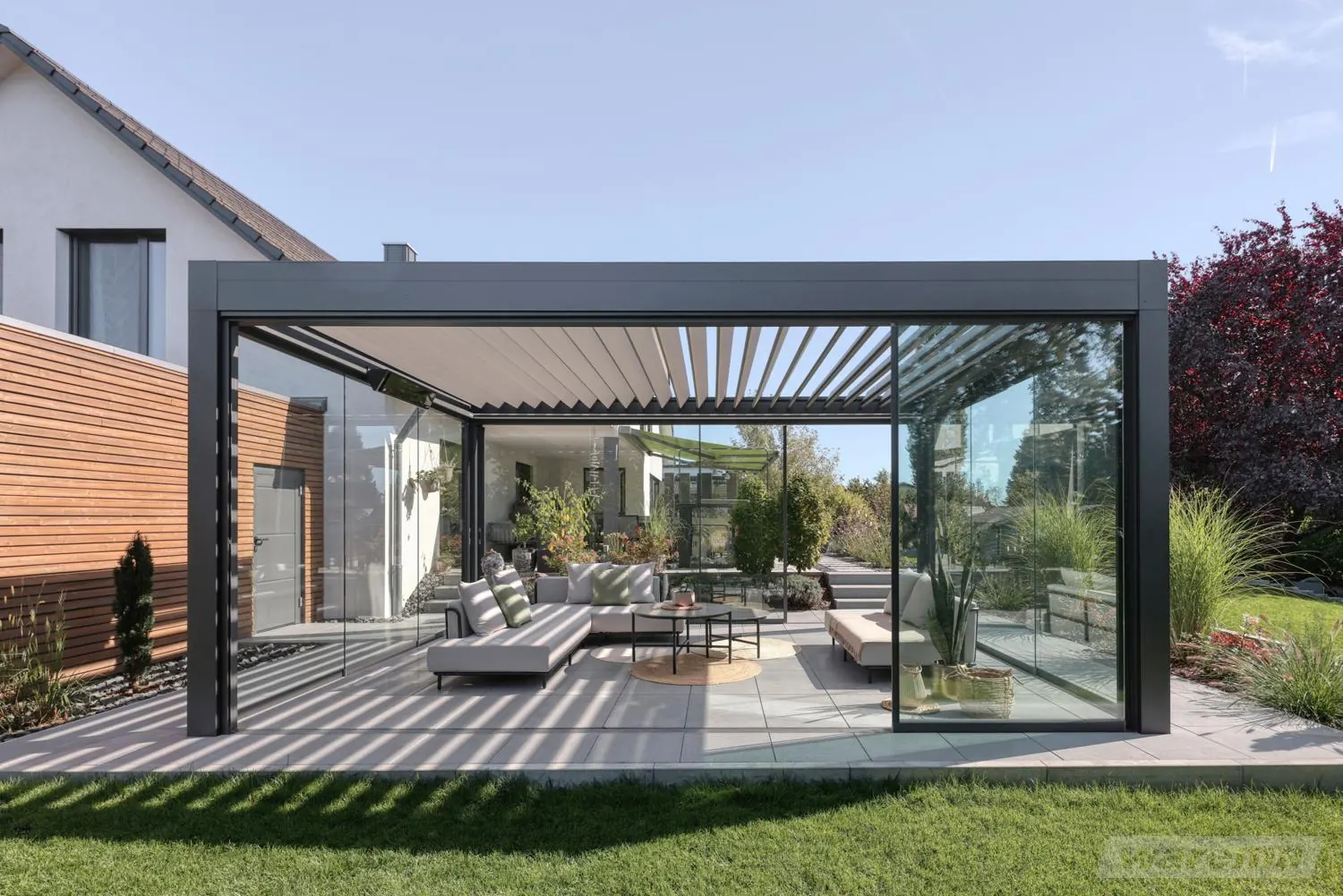 Lamellendach mit rahmenlosen Glasschiebewänden rundum, Lounge im Trockenen, über dem Sitzplatz geöffnete Lamellen
