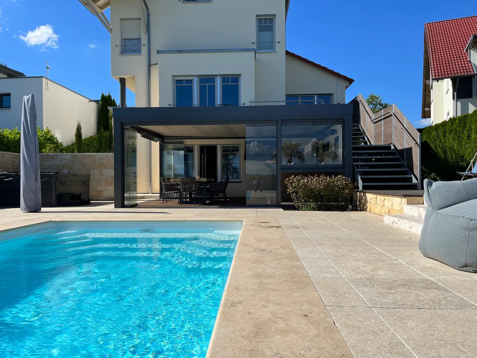 Terrassenüberdachung mit Glas-Schiebewänden auf drei Seiten neben einem Pool, dahinter das Wohnhaus