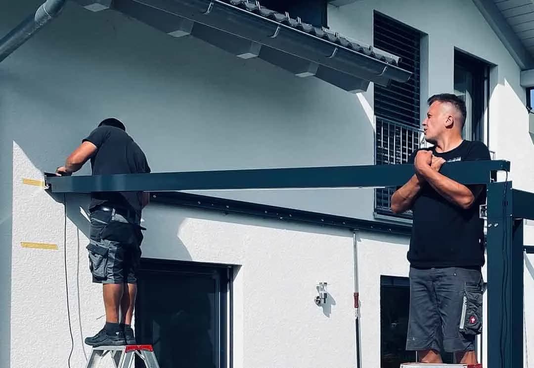 Zwei Monteure befestigen den Wandträger einer Terrassenüberdachung an der Hausfassade