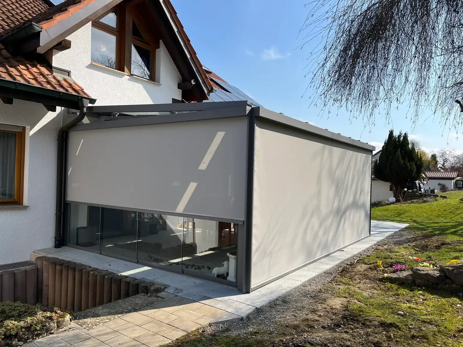 Der Glasanbau am Giebel des Hauses, an Front und Längsseite sind die beigen Senkrechtmarkisen heruntergelassen