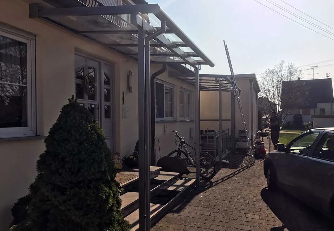 Blick an der Fassade entlang: vorn die Eingangsüberdachung über der Natursteintreppe, hinten entsteht das zweite Glasdach