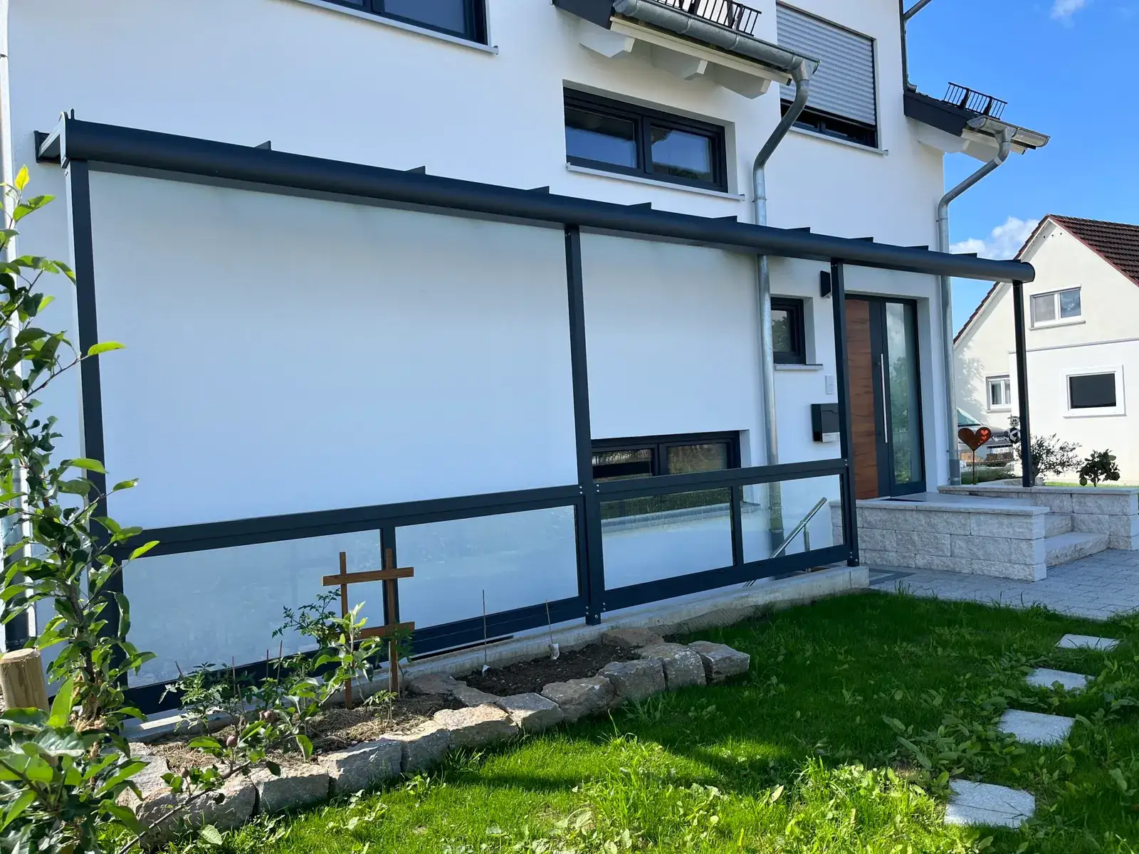 Die Längsseite der Überdachung: das dunkle Dach läuft an der weißen Fassade entlang, darunter die Glasbrüstung, im Hintergrund die Haustür