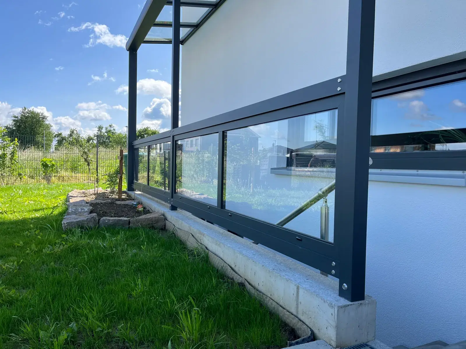 Detail der Stütze auf der Kante neben dem Rasen, daneben die Glasbrüstung, dahinter die weiße Hauswand