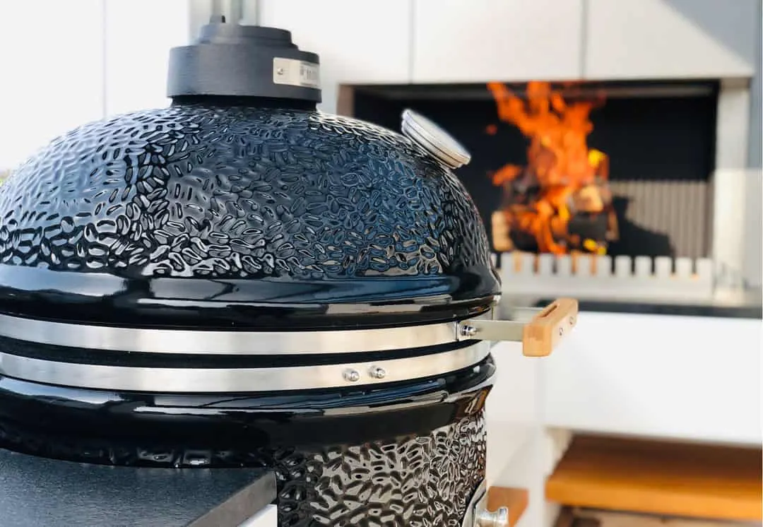 Schwarzer Keramikgrill im Vordergrund, dahinter brennt ein Feuer in der gemauerten Feuerstelle der Outdoor-Küche