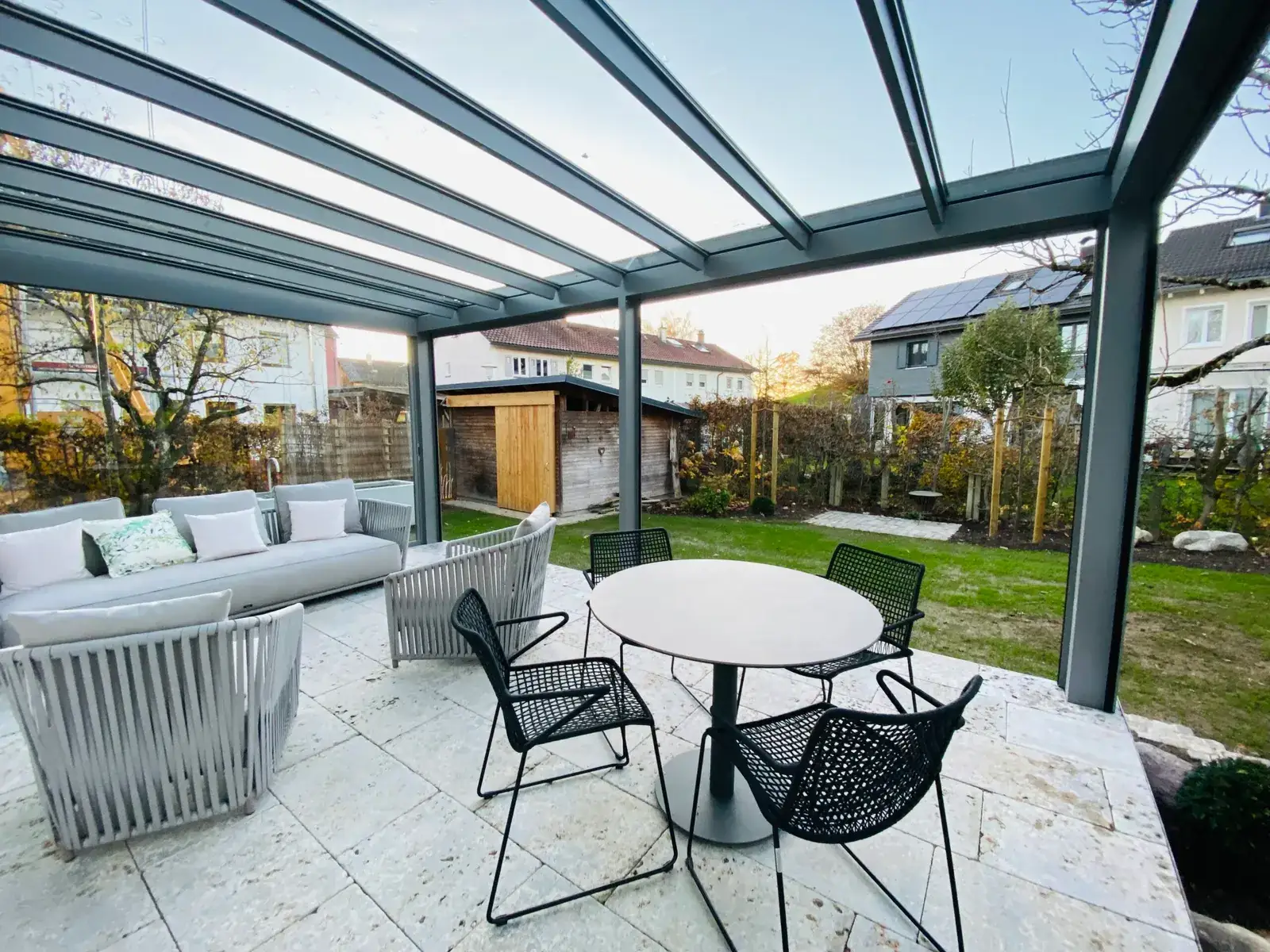 Sitzgruppe unter dem Glasdach mit rundem Tisch und schwarzen Stühlen, dahinter der frisch angelegte Garten