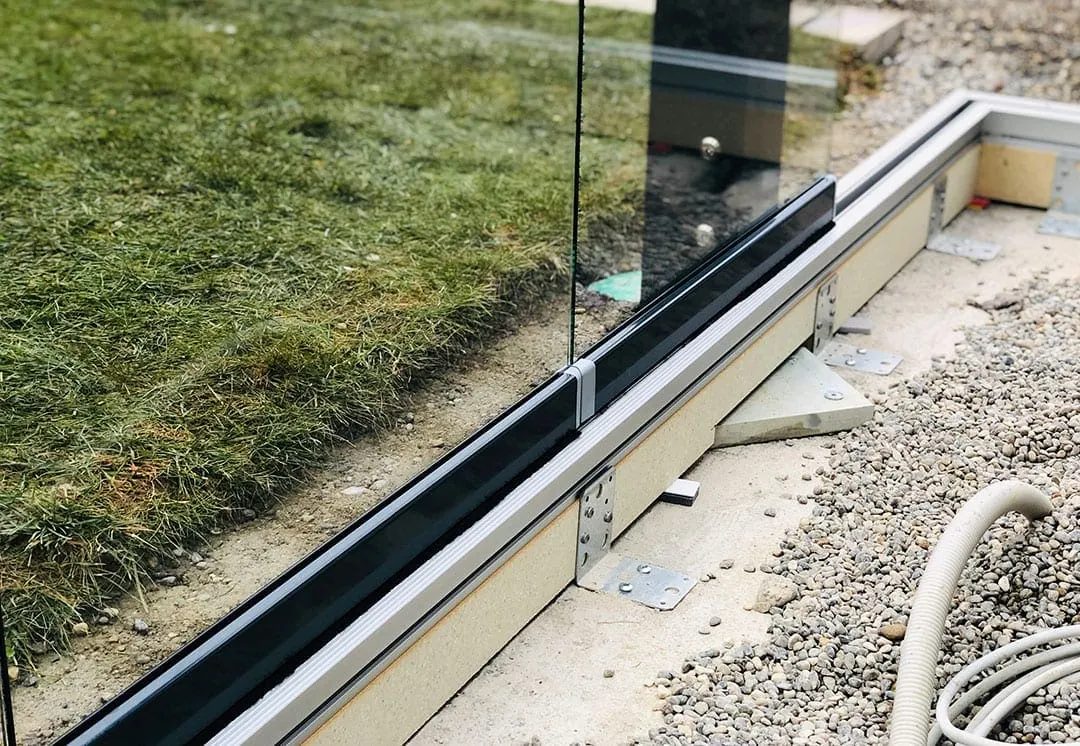 Detail der bodennahen Laufschiene: die rahmenlosen Glasflügel laufen in einer flachen Schiene, darunter verzinkte Winkelbleche und lose Platten im Kies, links der Rasen