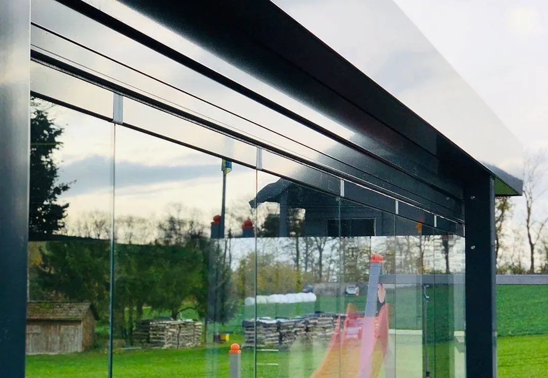 Blick von unten auf die Dachkante und die Glasflügel, dahinter die Wiese mit Holzstapel, kleiner Hütte und der roten Rutsche
