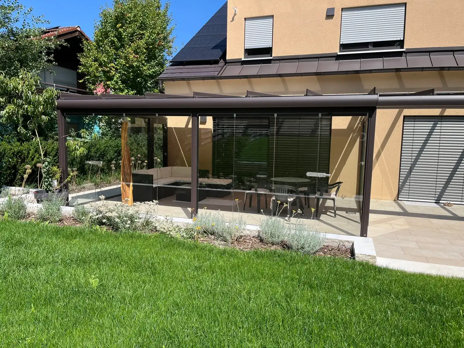Der geschlossene Kaltwintergarten vom Rasen aus: hinter den Glas-Schiebewänden eine helle Sitzecke und eine Holzskulptur, davor ein Staudenbeet