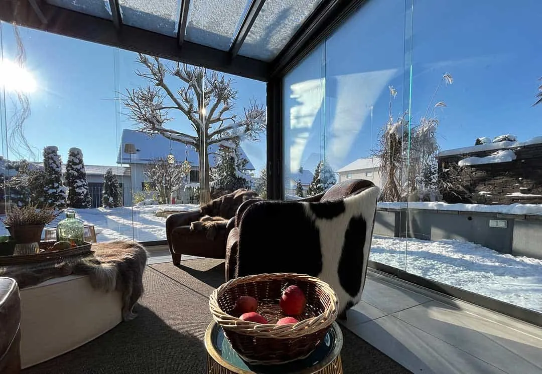 Blick aus dem Kaltwintergarten in den verschneiten Garten, auf dem niedrigen Tisch steht eine Korbschale mit roten Äpfeln