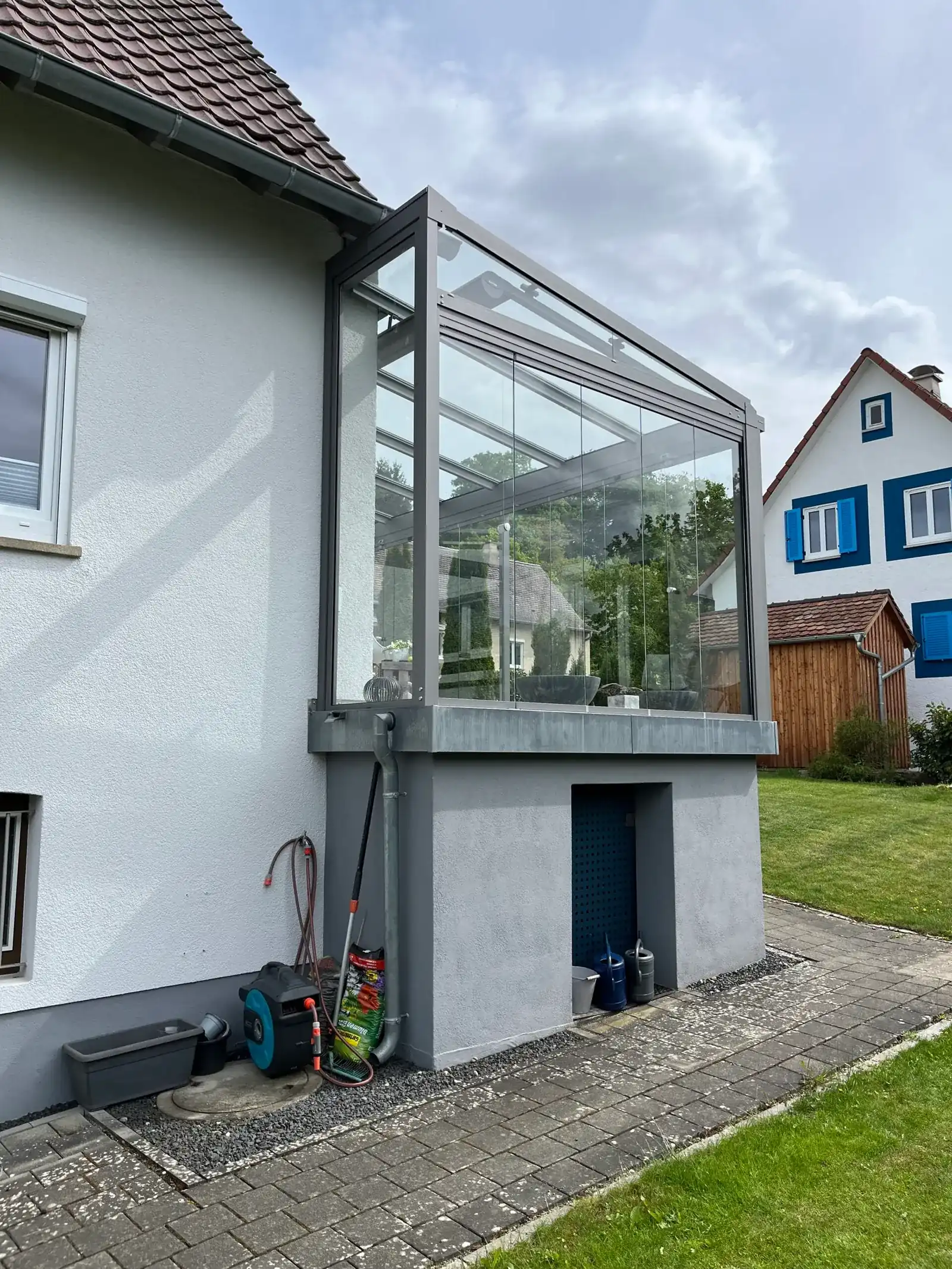 Der Kaltwintergarten von der Seite: Glasfront auf dem verputzten Sockel, darunter eine offene Nische mit einem Eimer und zwei Gießkannen