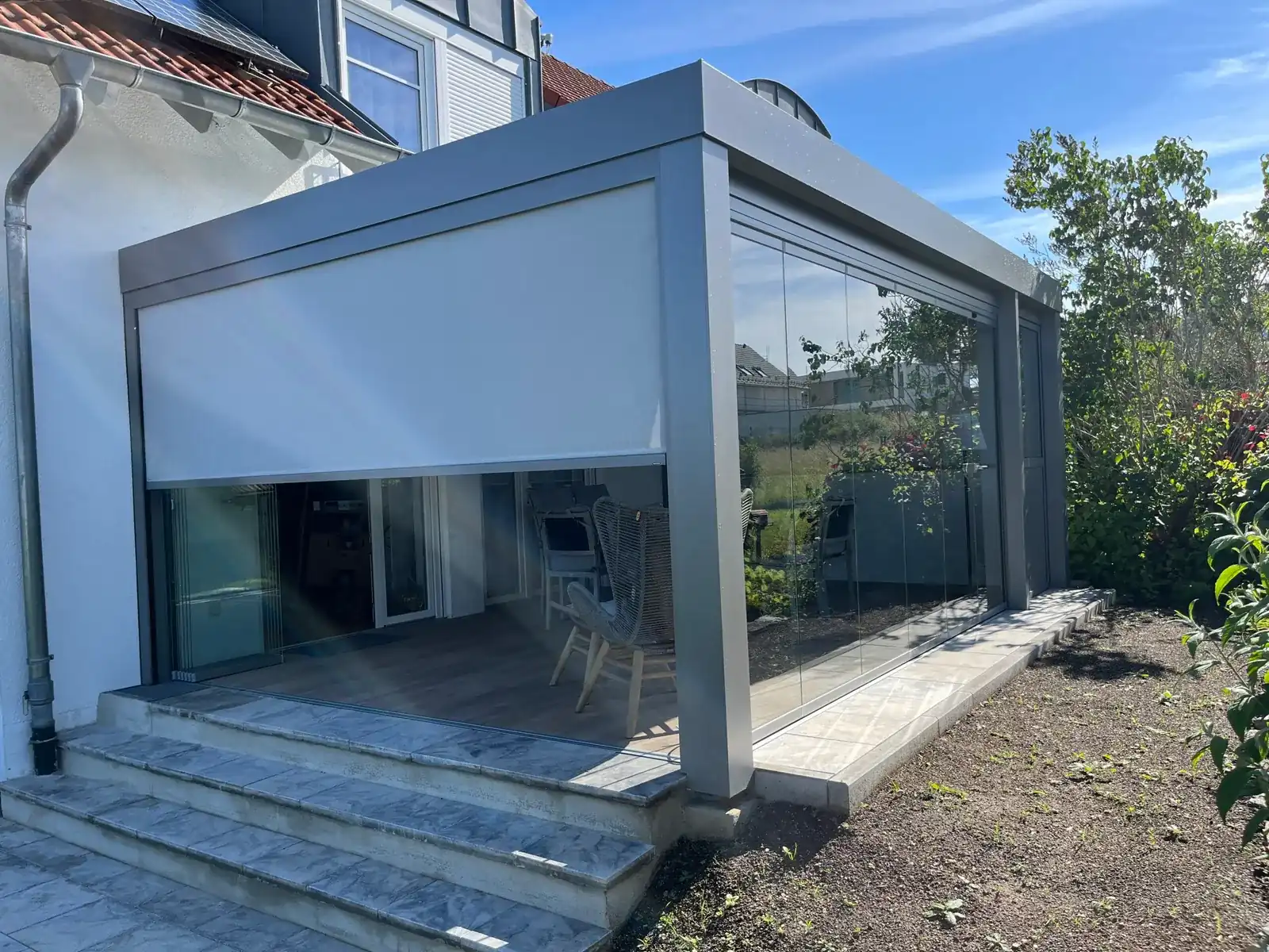 Das hellgraue Terrassendach von der Gartenecke aus, rechts die Glaselemente, links das halb heruntergelassene Tuch der Senkrechtmarkise, davor die Steinstufen