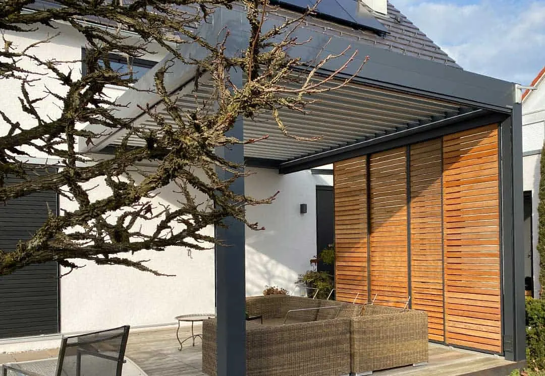 Der alte knorrige Baum vor dem anthrazitfarbenen Lamellendach, dahinter die Holzläden und Loungemöbel auf der Terrasse