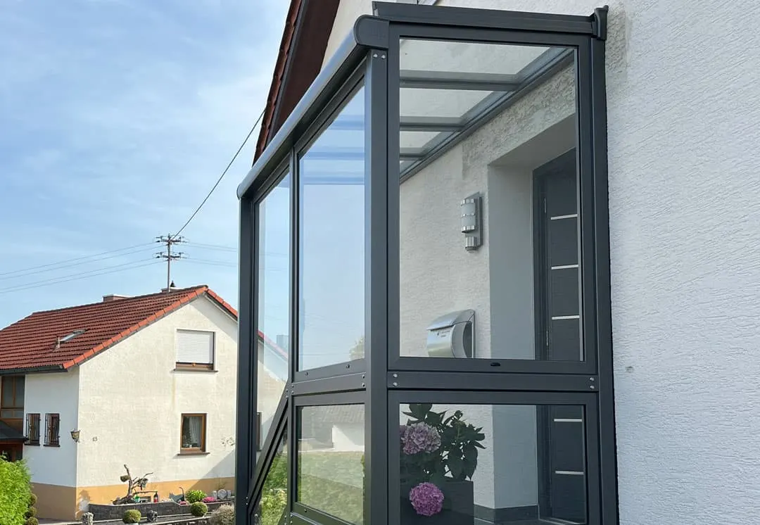 Der anthrazitfarbene Glasvorbau am weißen Haus, hinter dem Glas die Haustür, links das Nachbarhaus