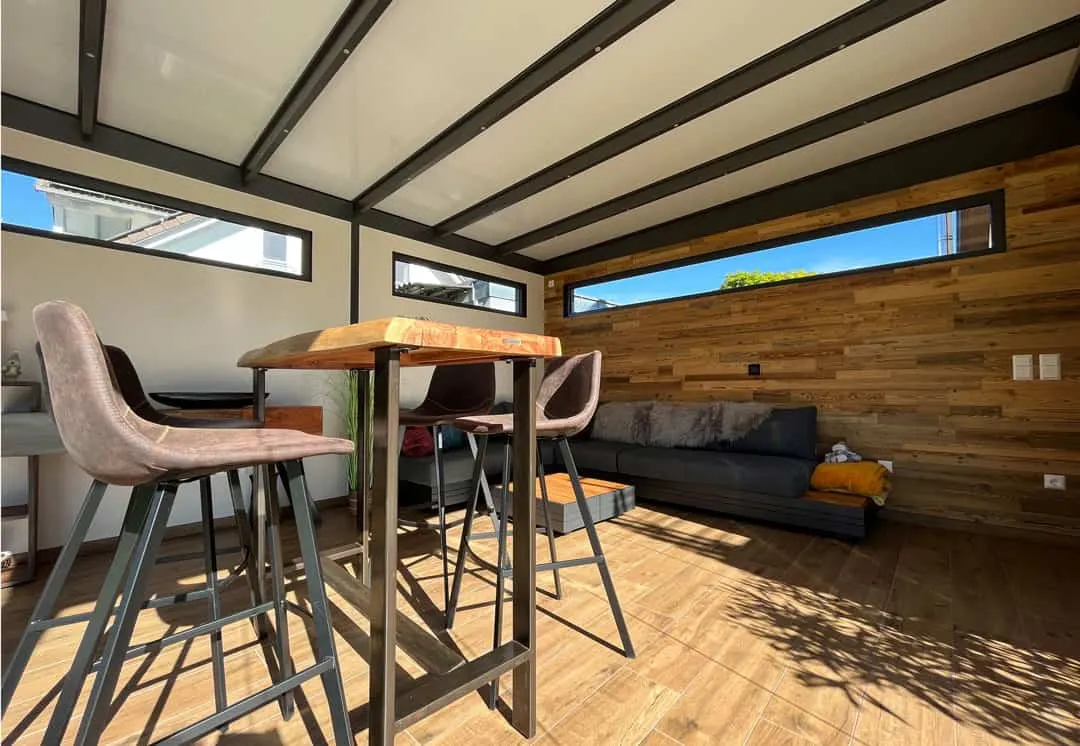 Innenansicht mit Bartisch aus Massivholz, Barhockern und Lounge-Sofa vor der holzverkleideten Wand mit schmalem Fensterband