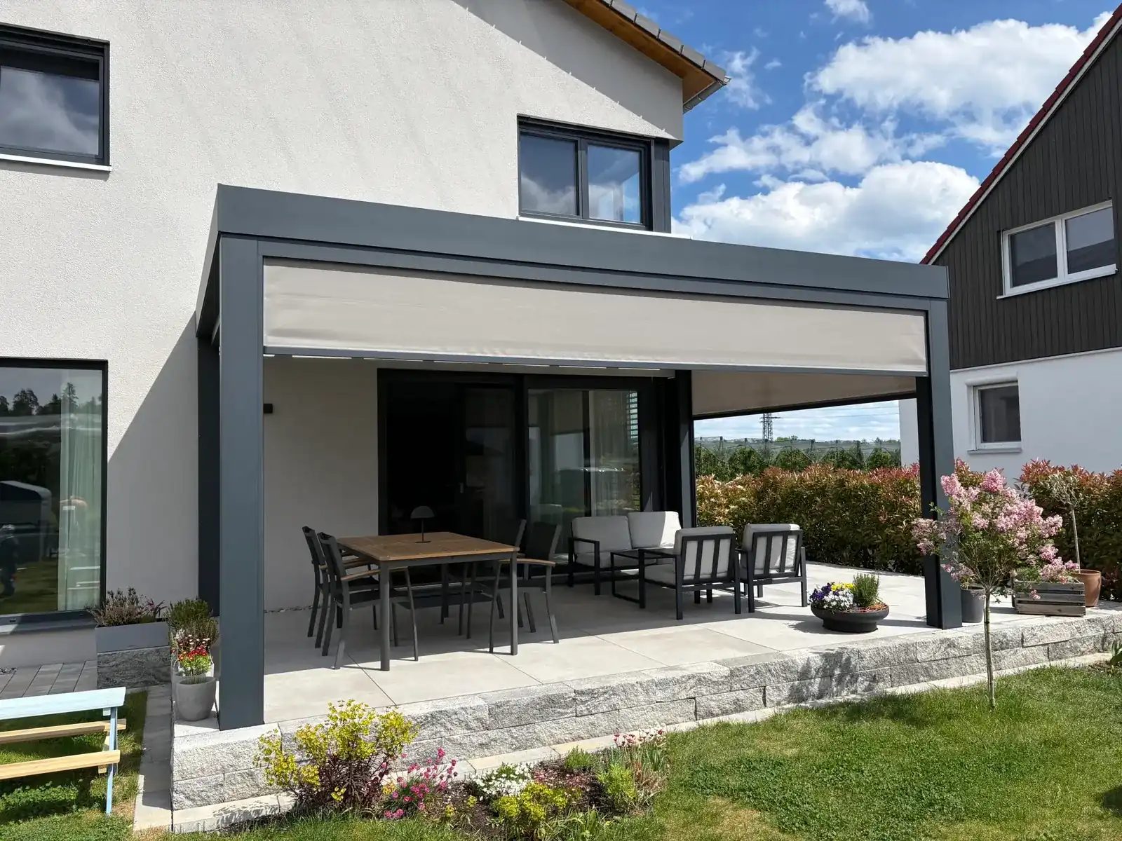 Front und Seite des Terrassendachs mit halb heruntergefahrenen Senkrechtmarkisen, die Terrasse liegt erhöht auf einer Granitsteinmauer