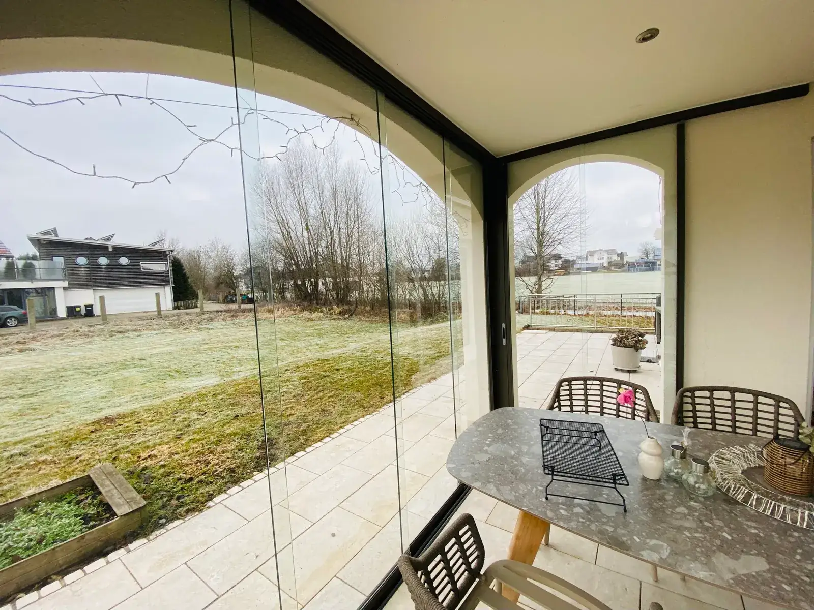 Blick aus der verglasten Terrasse über die Wiese auf Nachbarhäuser, links die Glasfelder im großen Rundbogen, rechts ein weiterer verglaster Bogen und der Esstisch