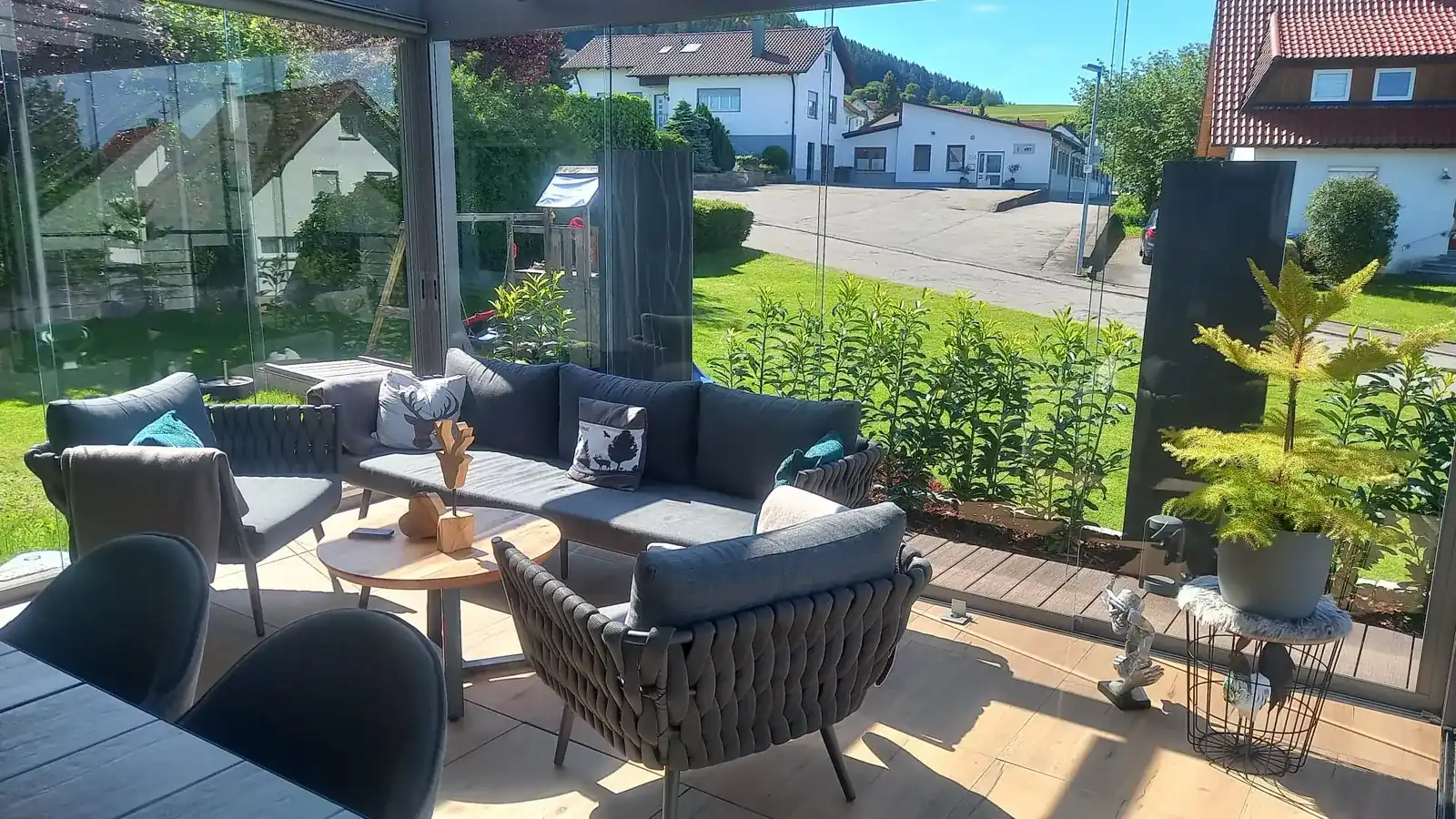 Blick von der Loungeecke unter dem Terrassendach über die Wiese auf die Nachbarhäuser und die bewaldeten Hügel dahinter