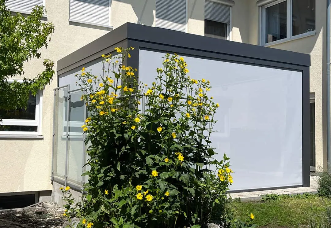 Die kubische Blende der Überdachung von der Seite, davor blühen hohe gelbe Stauden, an der Front ist die Senkrechtmarkise heruntergelassen