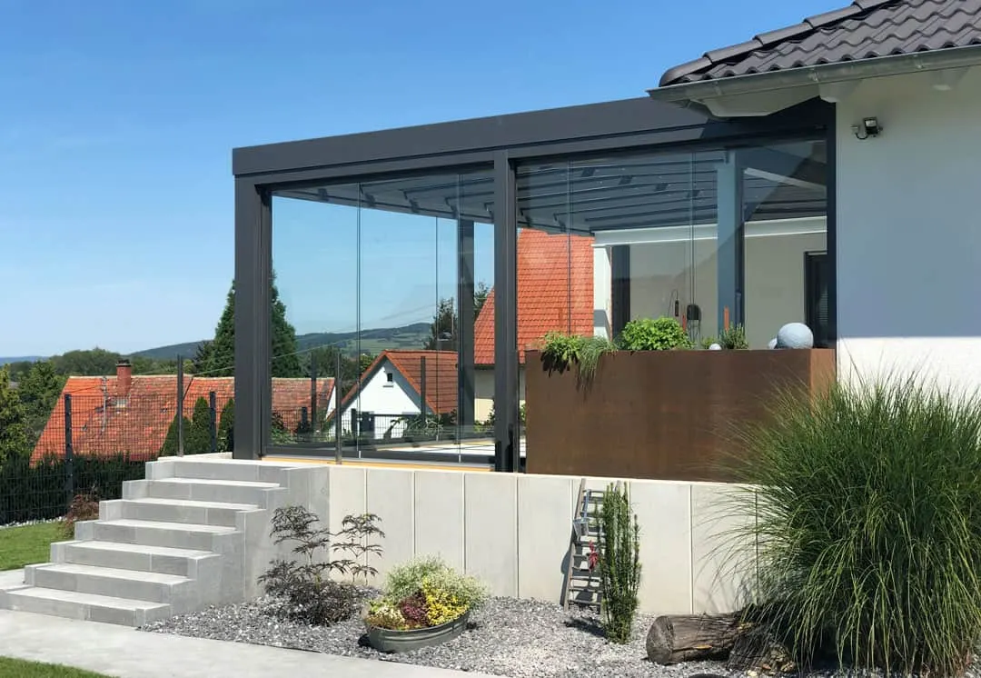 Das kubische Terrassendach von der Seite: Glas-Schiebewände über dem Betonsockel, ein Cortenstahl-Pflanzkasten und der Blick über die Dächer der Nachbarschaft