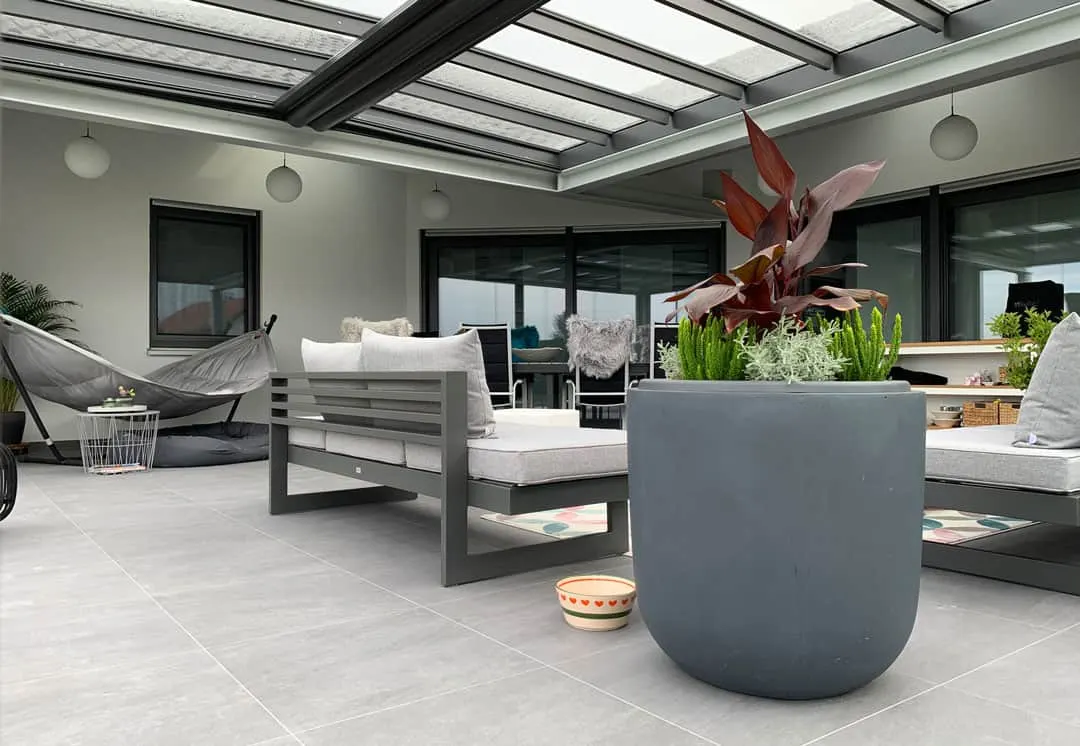 Unter dem Glasdach: Lounge-Sofa mit hellen Kissen, Hängematte und große Pflanzkübel auf grauen Bodenplatten
