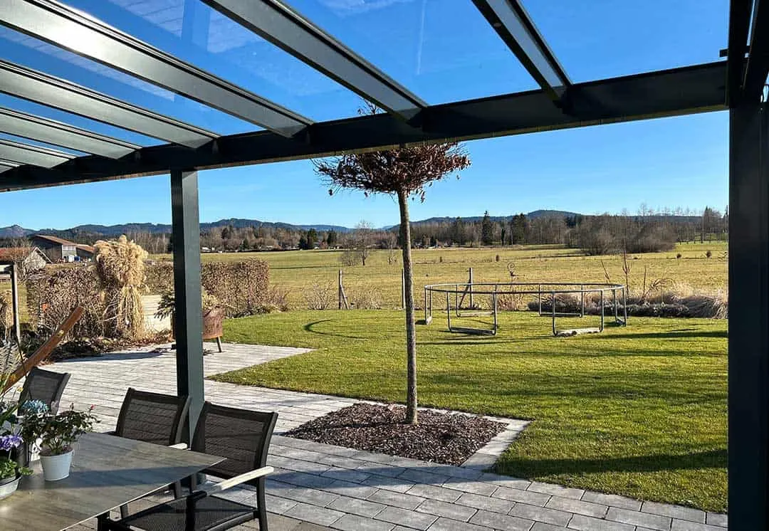 Blick von der Terrasse unter dem Glasdach über den Rasen auf offene Wiesen und bewaldete Hügel