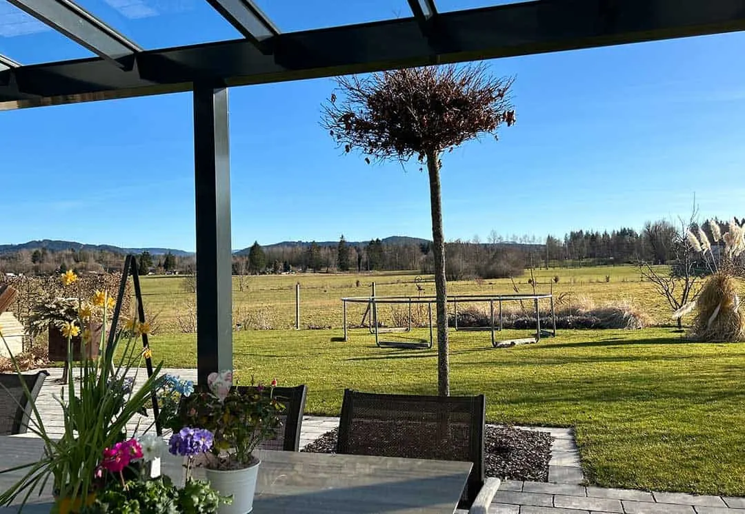 Frühlingsblumen auf dem Terrassentisch unter dem Glasdach, dahinter Wiesen bis zu den Hügeln