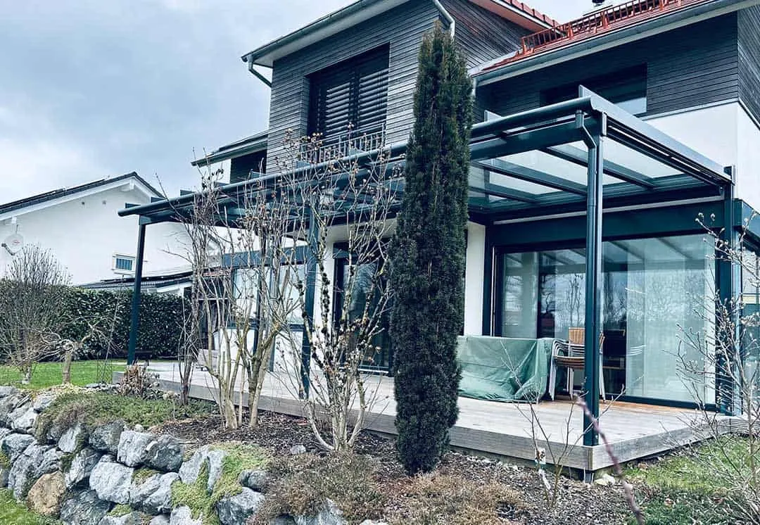 Die Ecke des Terrassendachs mit raumhoher Seitenverglasung, davor eine schmale Säulenkonifere