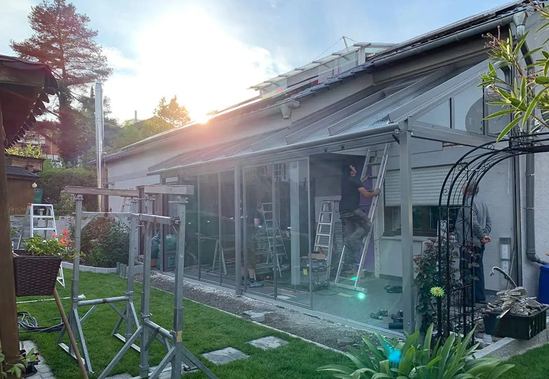 Abendsonne über dem Hausdach, im Glasanbau ein Monteur auf der Leiter, links davon eine zweite Person, rechts außen eine dritte im grauen Kapuzenpulli