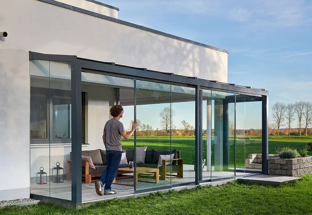 Ein Mann schiebt die Glaswand des Terrassendachs am weißen Flachdachhaus auf, dahinter weite Felder
