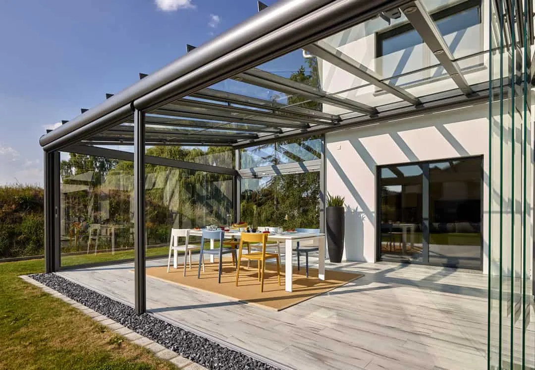 Das dunkelgraue Terrassendach mit Glas-Schiebewänden reicht über die Hausecke des weißen Wohnhauses hinaus