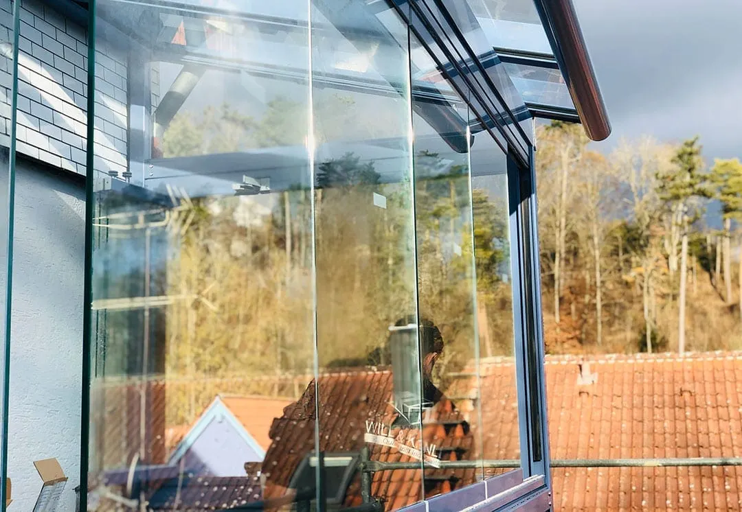 Blick an der Glasfront entlang über die Ziegeldächer der Nachbarhäuser zum bewaldeten Hang, auf der Scheibe klebt das Firmenschild von Wild und Kienle