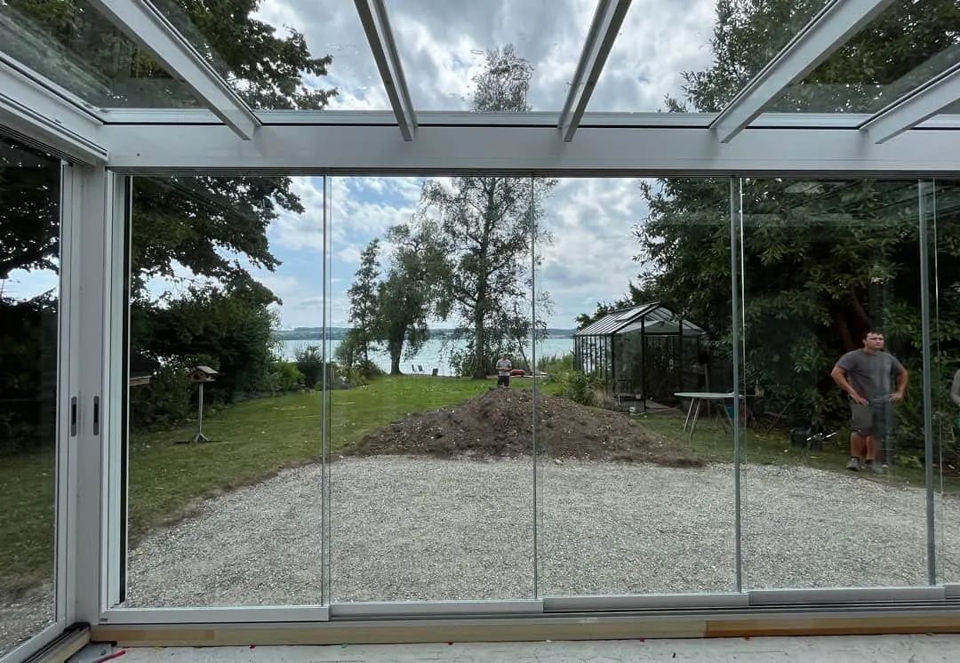 Blick aus dem Glasraum über den Garten zum See, rechts steht ein Mann in grauem Shirt, im Garten arbeitet eine zweite Person