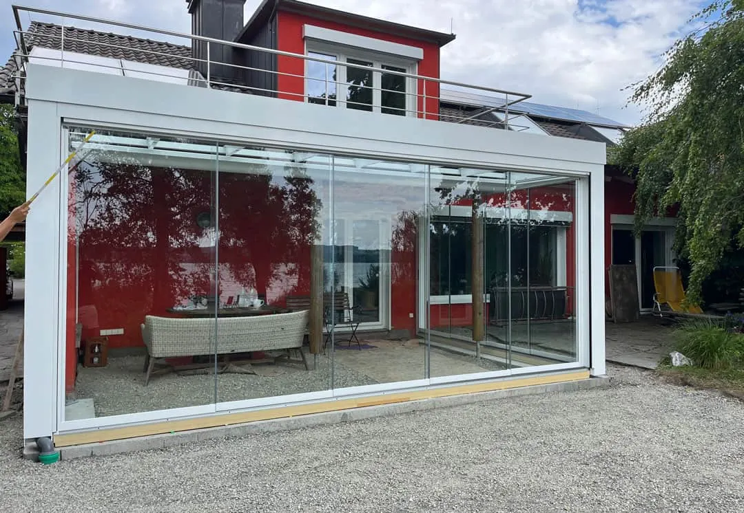 Vorderseite des Glashauses am roten Haus, am linken Bildrand zieht eine Hand einen Wischer über die Scheibe
