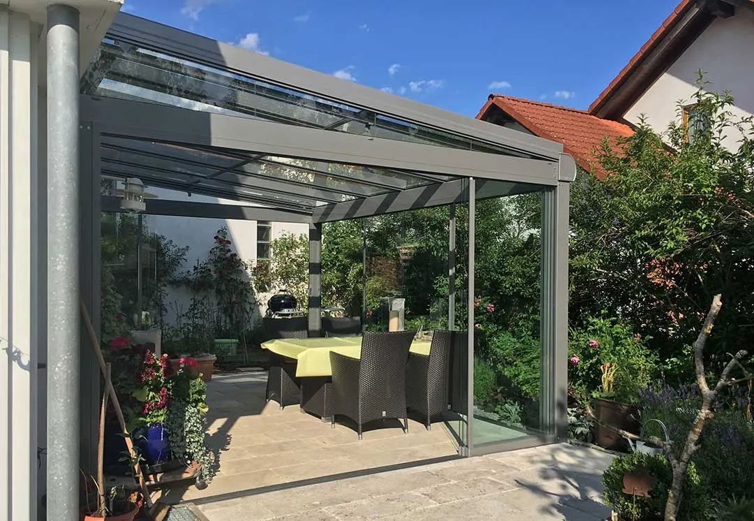 Die geöffnete Front der Überdachung mit zur Seite geschobenen Glaselementen, innen der Tisch mit gelber Decke