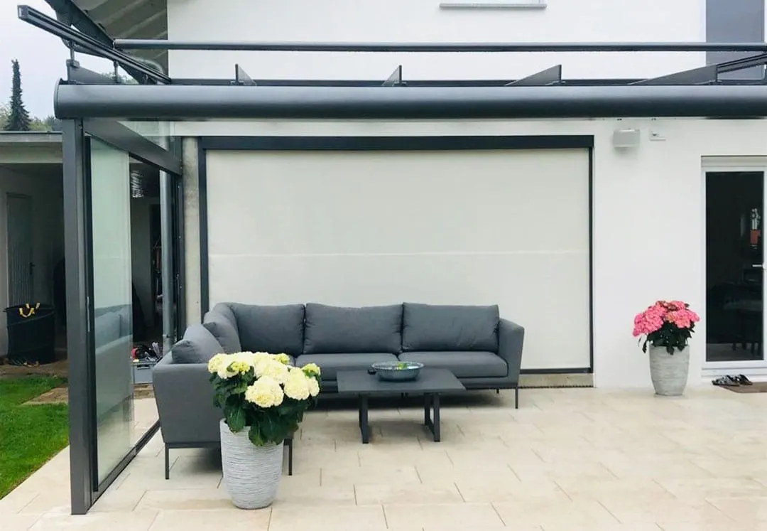 Die heruntergefahrene helle Senkrechtmarkise hinter dem grauen Ecksofa, rechts eine Hortensie im Kübel