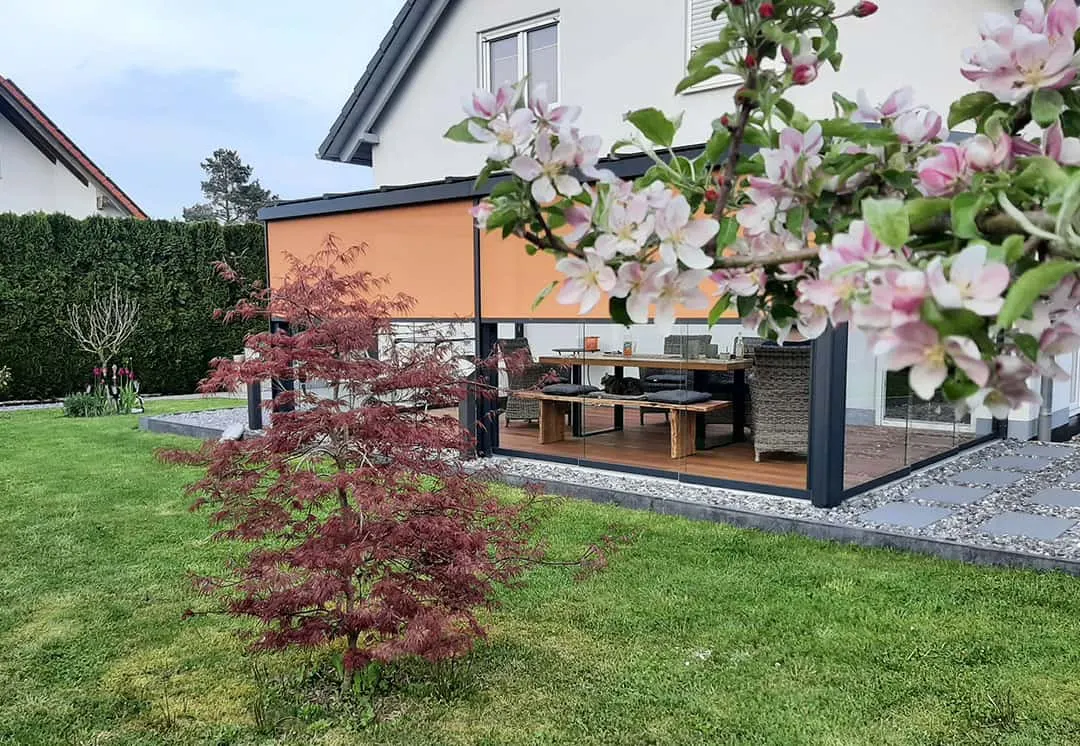 Das Terrassendach vom Rasen aus mit halb heruntergelassener oranger Senkrechtmarkise, davor ein roter Fächerahorn
