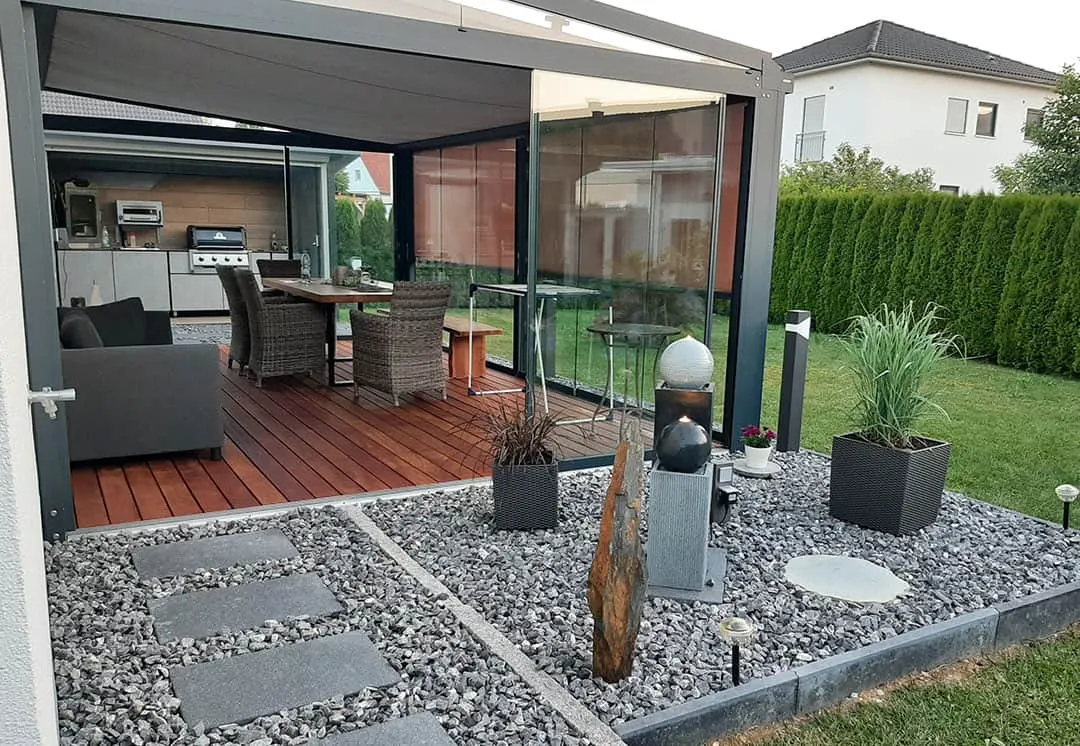 Blick von der Kiesfläche unter das Dach: Küchenzeile mit Grill, Esstisch mit Geflechtsesseln, Sofa und rechts ein Wasserspiel