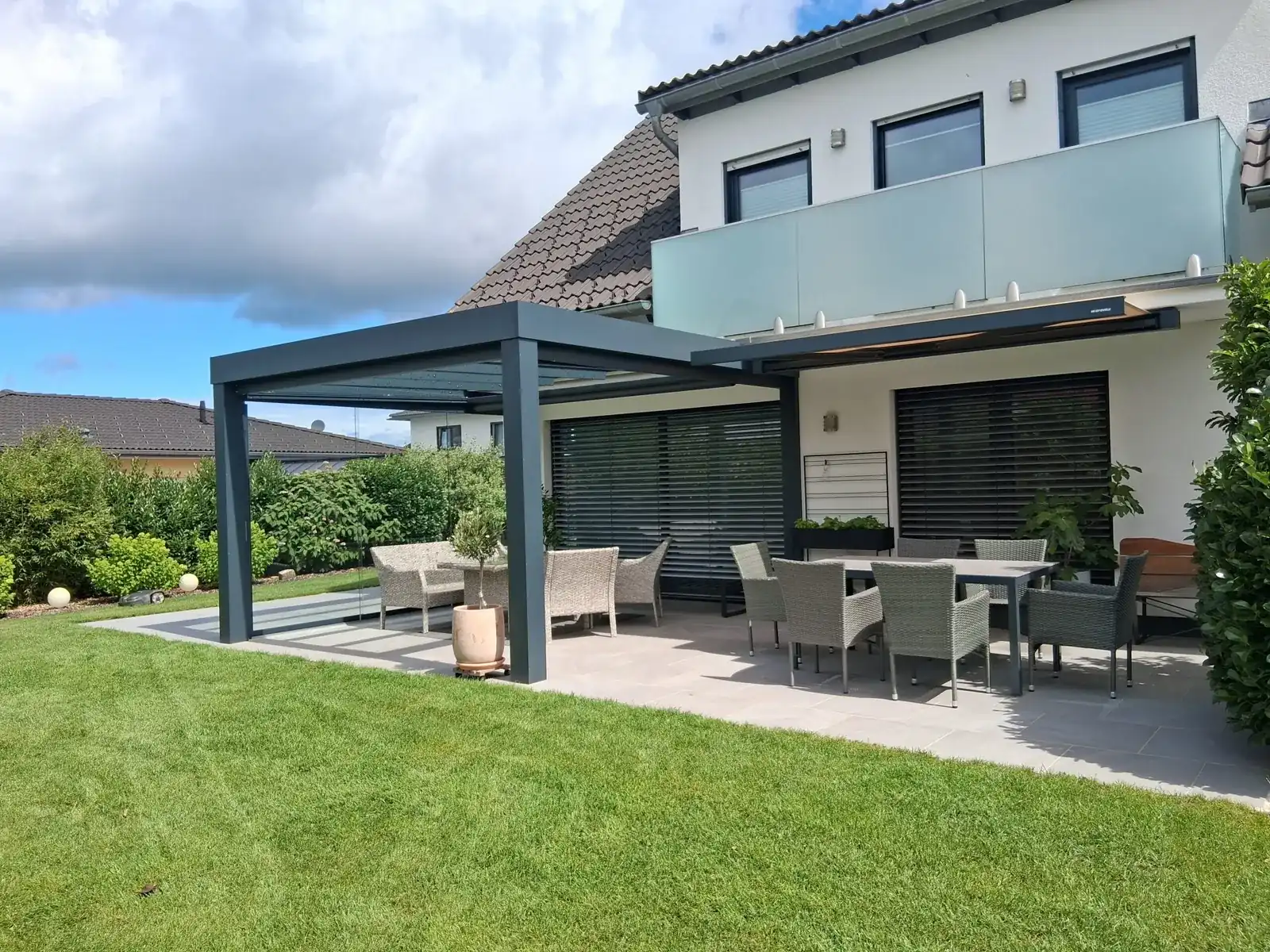 Die Terrasse mit zwei Dächern: links das Glasdach mit gerader Kante, rechts die ausgefahrene sandfarbene Markise überm Esstisch