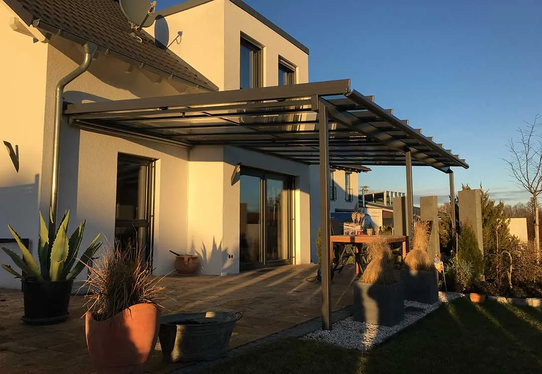 Das dunkelgraue Terrassendach mit überstehender Glaskante in tiefer Wintersonne, davor Kübelpflanzen auf der Terrasse
