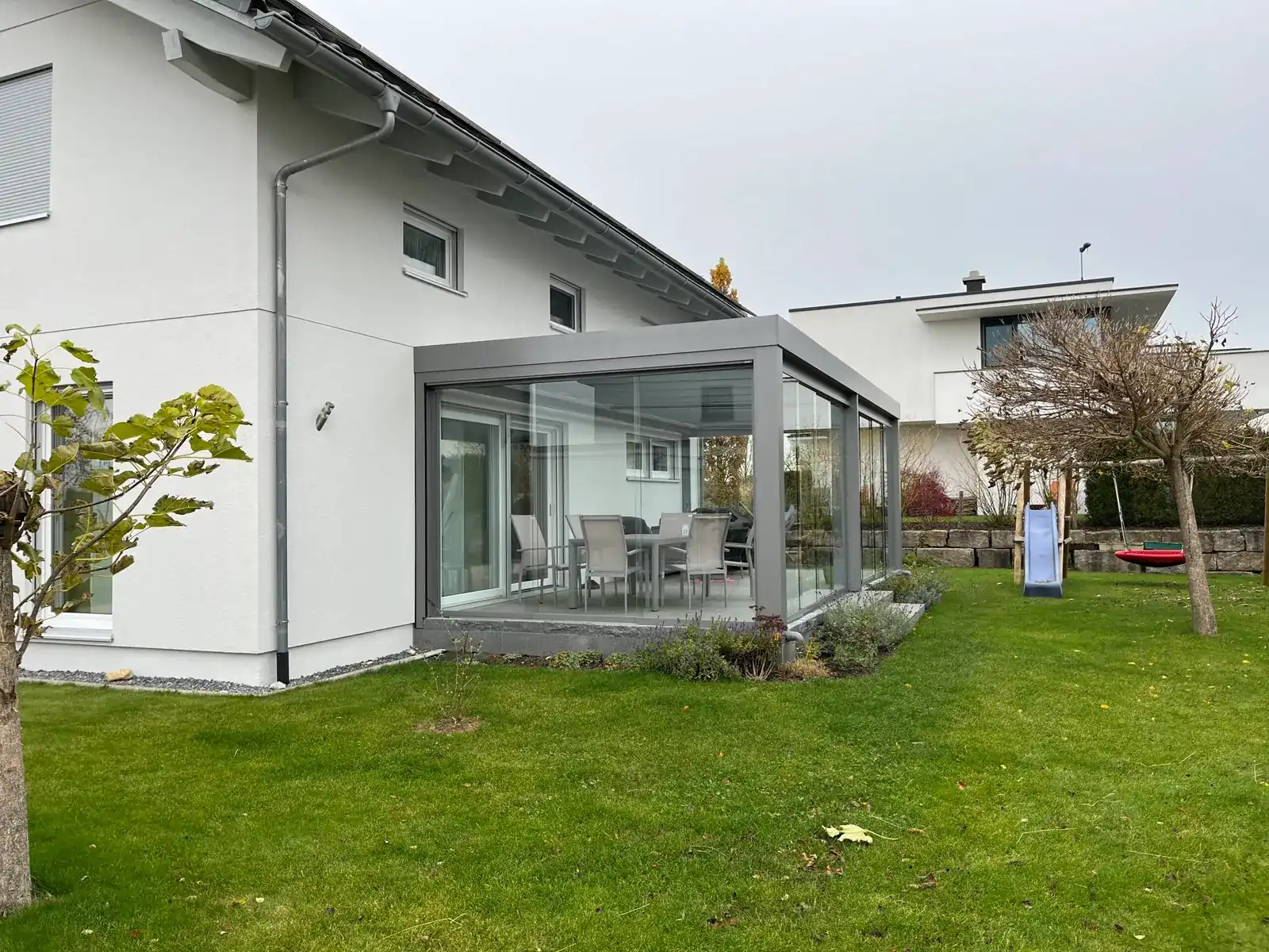 Der graue Glasanbau in der Ecke des Einfamilienhauses, dahinter der Rasen mit Rutsche und Nestschaukel