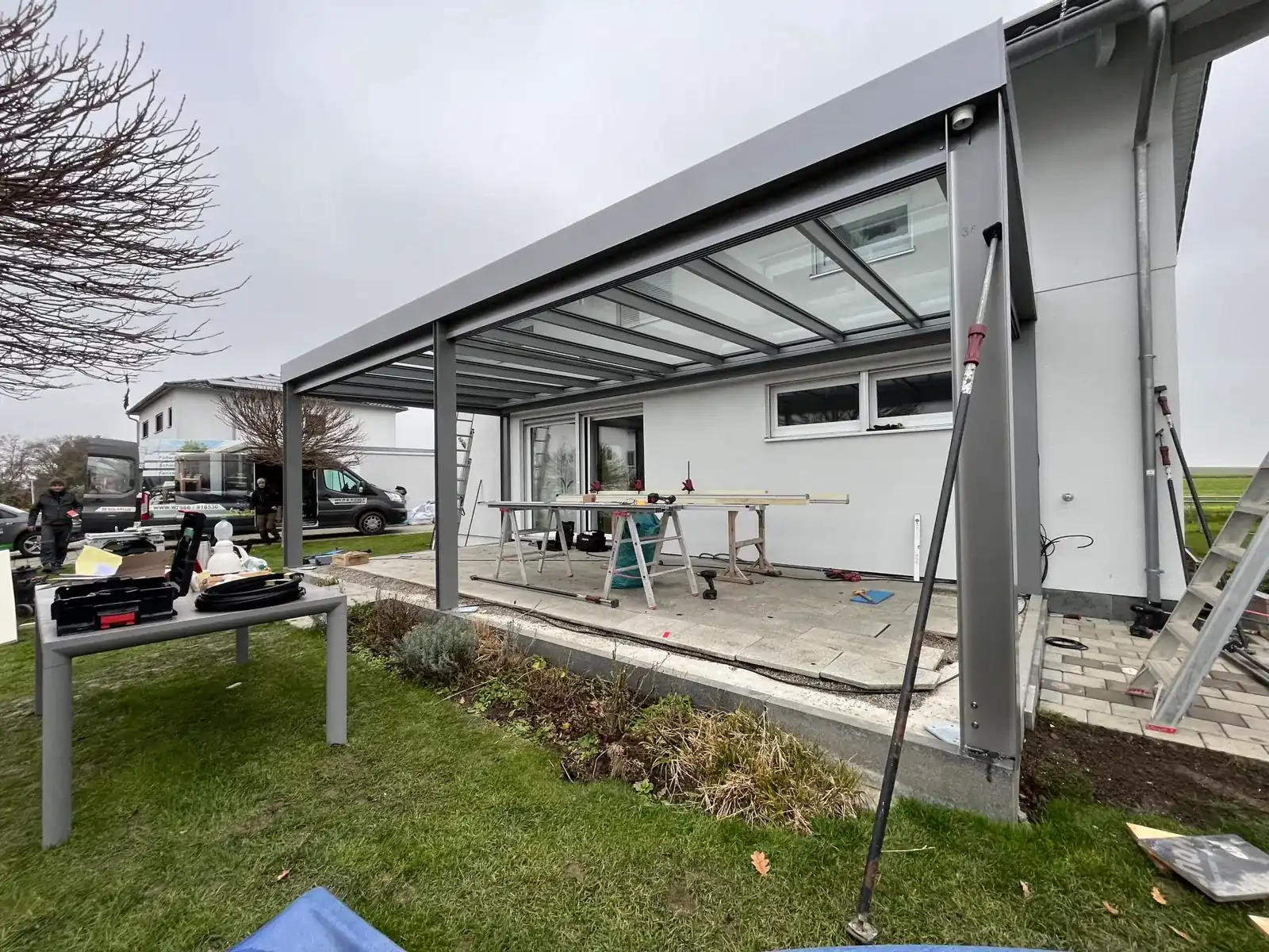 Montage: der graue Rahmen mit fertigem Glasdach steht noch ohne Seitenwände, zwei Monteure neben dem Transporter, Werkzeug auf den Terrassenplatten