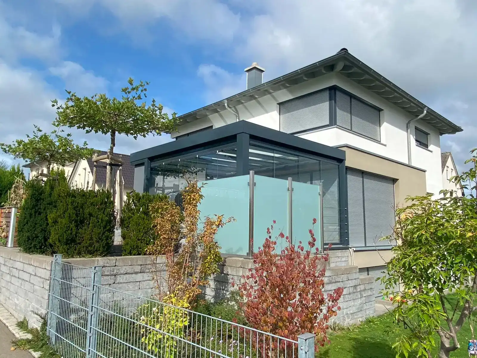 Das Haus von der Straße aus: der anthrazitfarbene Glasanbau an der Hausseite zur Straße, davor die Mauer und ein Sichtschutz aus mattem Glas