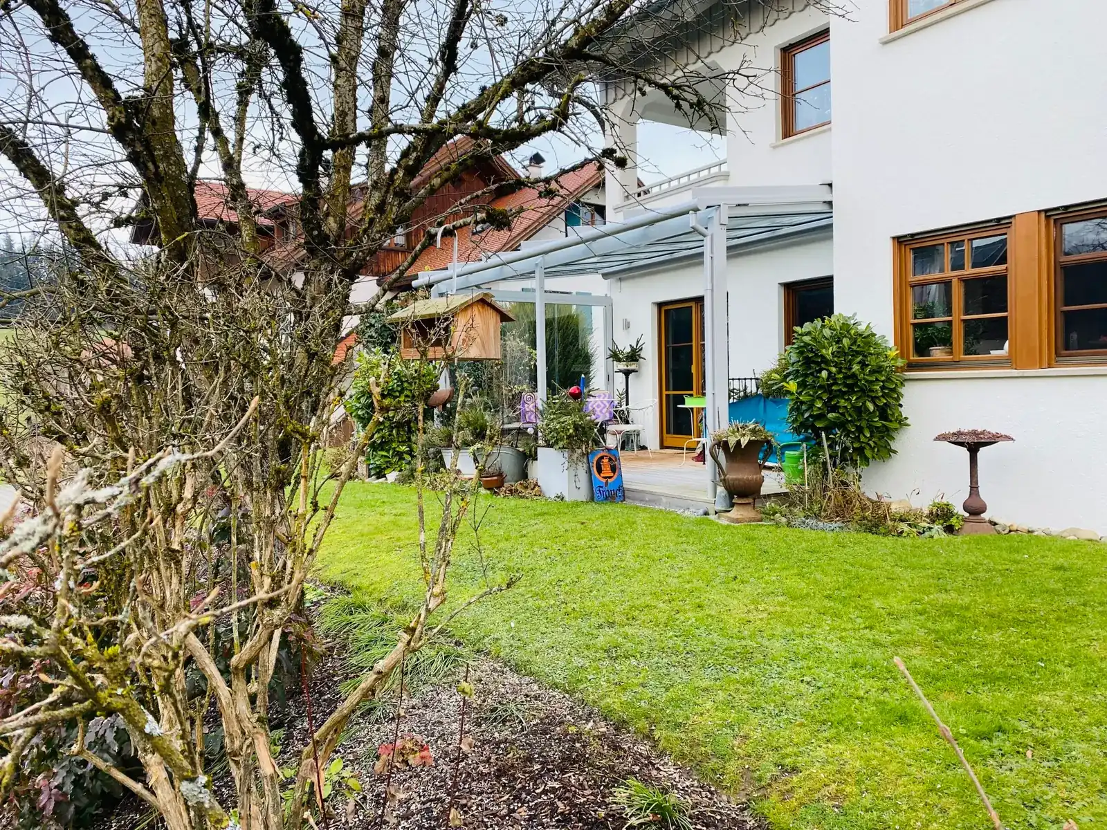 Blick über den Rasen auf das helle Terrassendach an der weißen Hausecke, im Vordergrund ein Vogelhaus in einem kahlen Baum