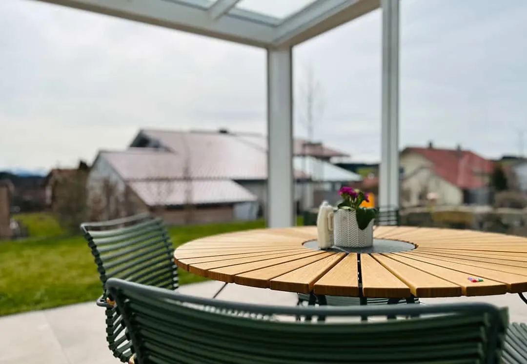 Runder Holztisch mit Kanne und kleinem Blumentopf unter dem Dach, dahinter der Blick über die Dächer der Nachbarhäuser