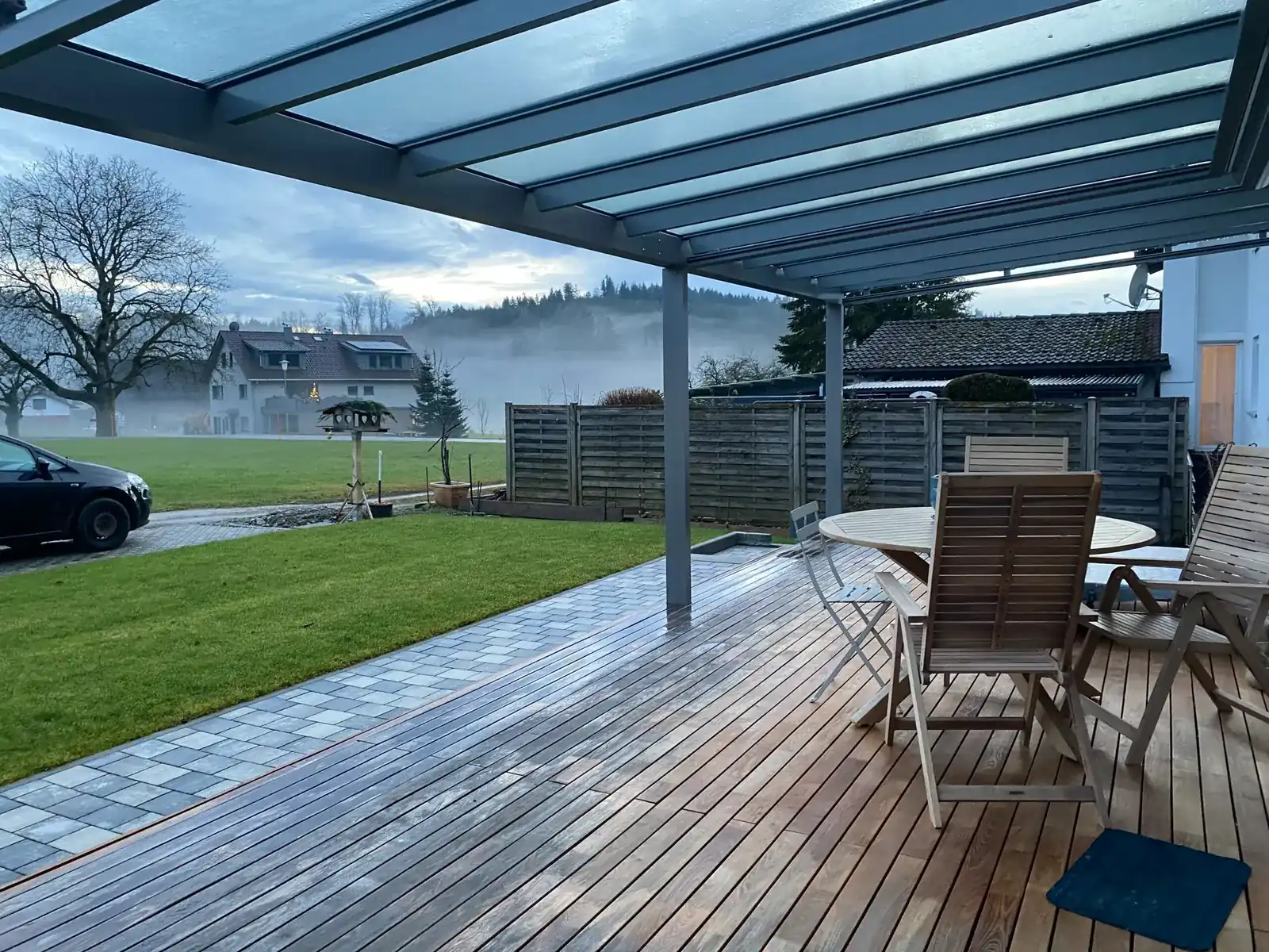 Blick von der Holzterrasse unter dem grauen Glasdach hinaus auf Rasen, Nachbarhäuser und einen bewaldeten Hügel, über den Wiesen ein Nebelband