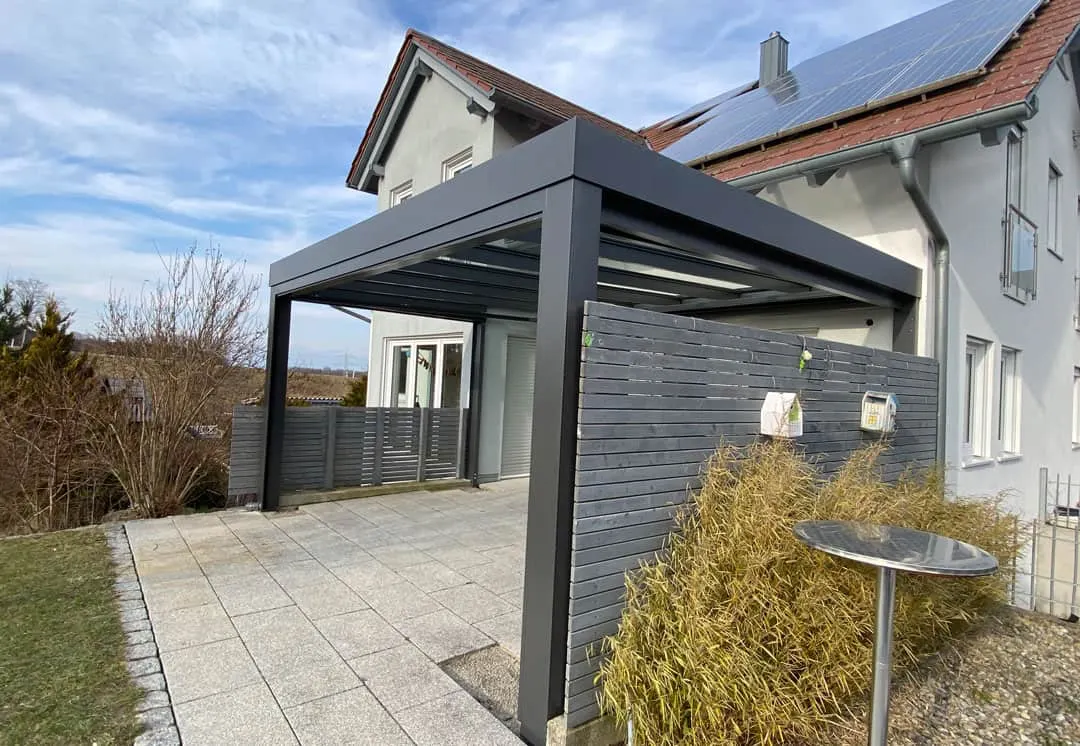Das Terrassendach von der Straßenseite: eine gerade graue Blende über der Lattenwand mit zwei kleinen Kästen, dahinter das Wohnhaus